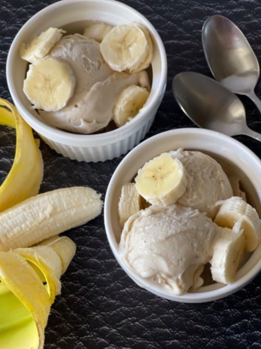two white bowls of frozen yogurt with banana sliced on top and on the side with two spoons