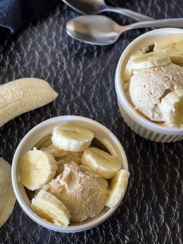 two white dishes with frozen yogurt and sliced bananas with two spoons in background and peeled banana in foreground