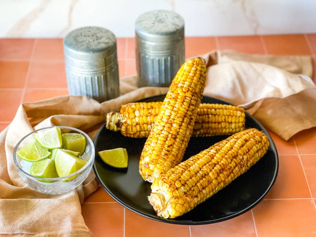 four air fryer corn on the cob on black plate with lime on side and salt and pepper shaker in the background