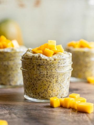 three jars of mango chia pudding with mangoes in the background in mango chunks on top