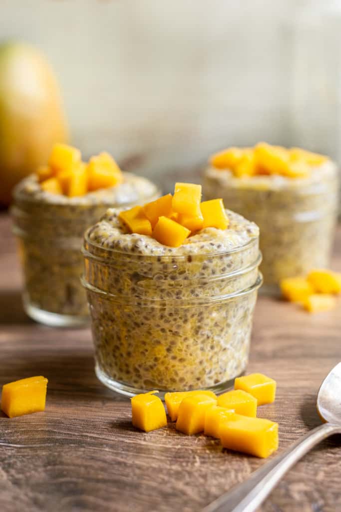 three jars of mango chia pudding with mango chunks on top and spoon on the side
