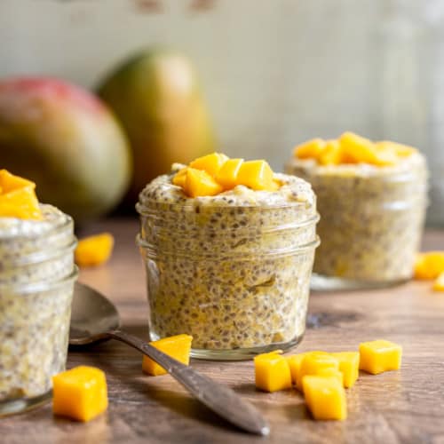three jars of mango chia pudding with mangoes in the background in mango chunks on top and spoon on the side