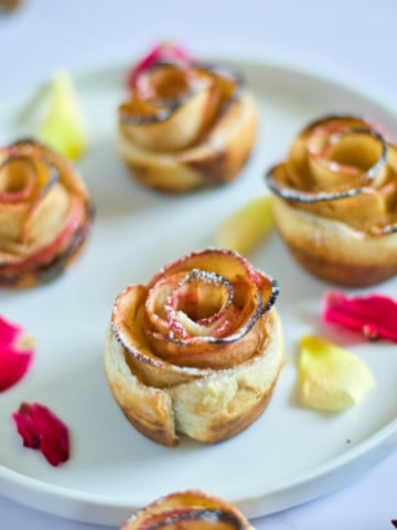 apple roses on white plate surrounded by cut apples, cinnamon sticks, roses, and wholee apple