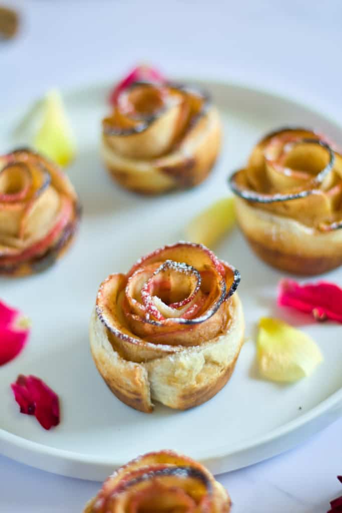 apple roses on white plate surrounded by cut apples, cinnamon sticks, roses, and wholee apple