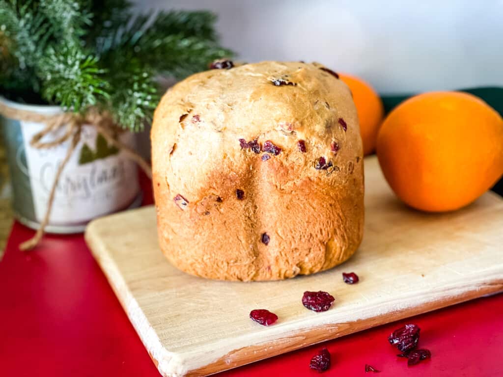 loaf of orange cranberry bread on wooden cutting board with dried cranberries in foreground, two oranges and christmas greenery in background