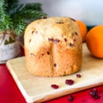 loaf of orange cranberry bread on wooden cutting board with dried cranberries in foreground, two oranges and christmas greenery in background