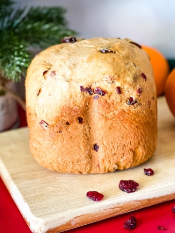 loaf of orange cranberry bread on wooden cutting board with dried cranberries in foreground, two oranges and christmas greenery in background