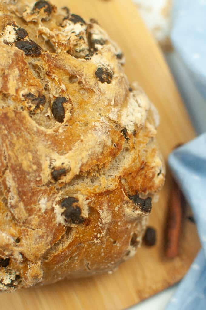 a round loaf of no-knead cinnamon raisin bread on a wooden cutting board with a blue cloth