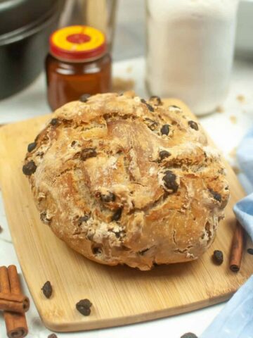 a round loaf of no-knead cinnamon raisin bread on a wooden cutting board with a blue cloth with raisins, cinnamon sticks, a jar of yeast and jar of flour in the background