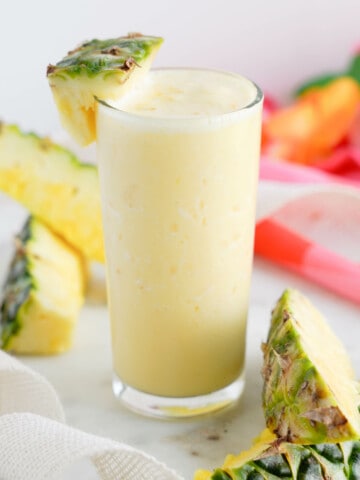 glass of orange pineapple smoothie with cut pineapple chunks to garnish. And orange and pink background.