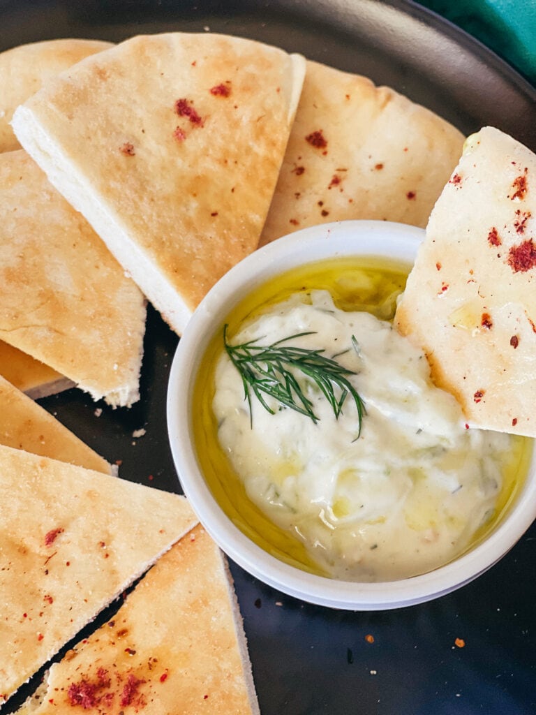 dipping a pita bread triangle into the vegan tzatziki. The sauce has a generous drizzle of oli oil and dill on top. the pita has a sprinkling of sumac