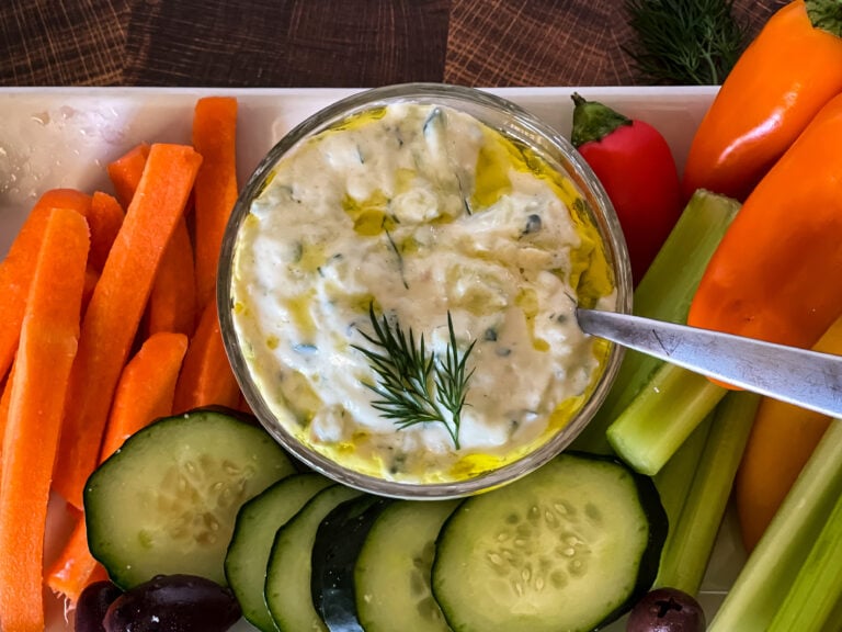 small bowl of vegan tzatziki sauce with plate of carrots, cucumbers, celery, olives, and peppers