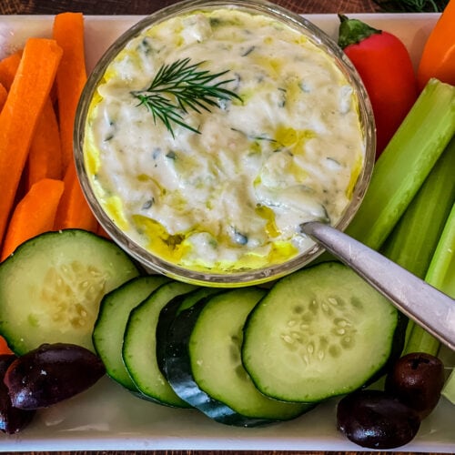 small bowl of vegan tzatziki sauce with plate of carrots, cucumbers, celery, olives, and peppers