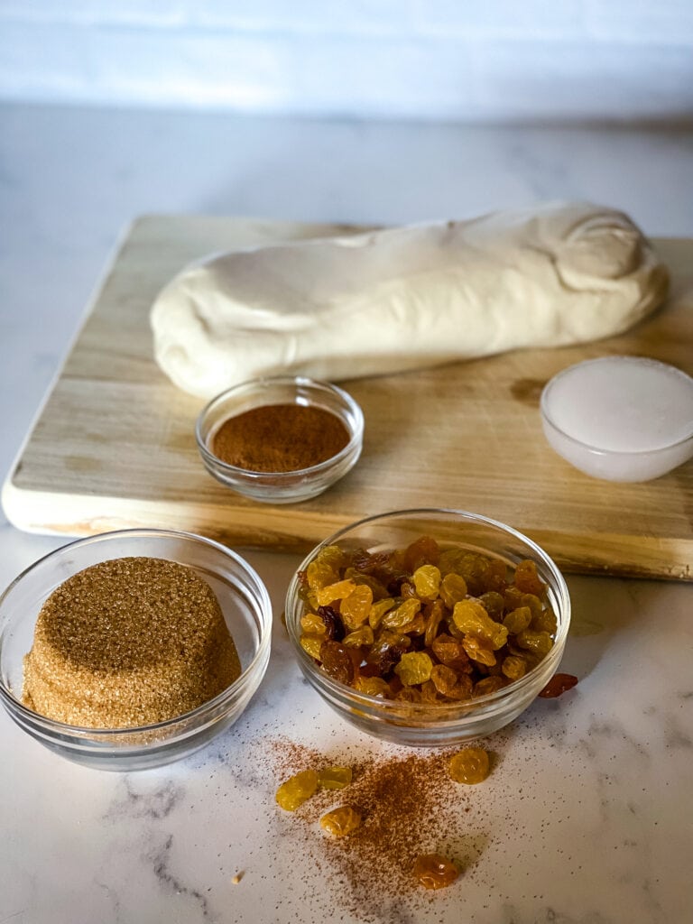 Ingredients needed for cinnamon swirl bread including bake and serve bread dough, cinnamon, coconut oil,c cinnamon, brown sugar, and golden raisins.