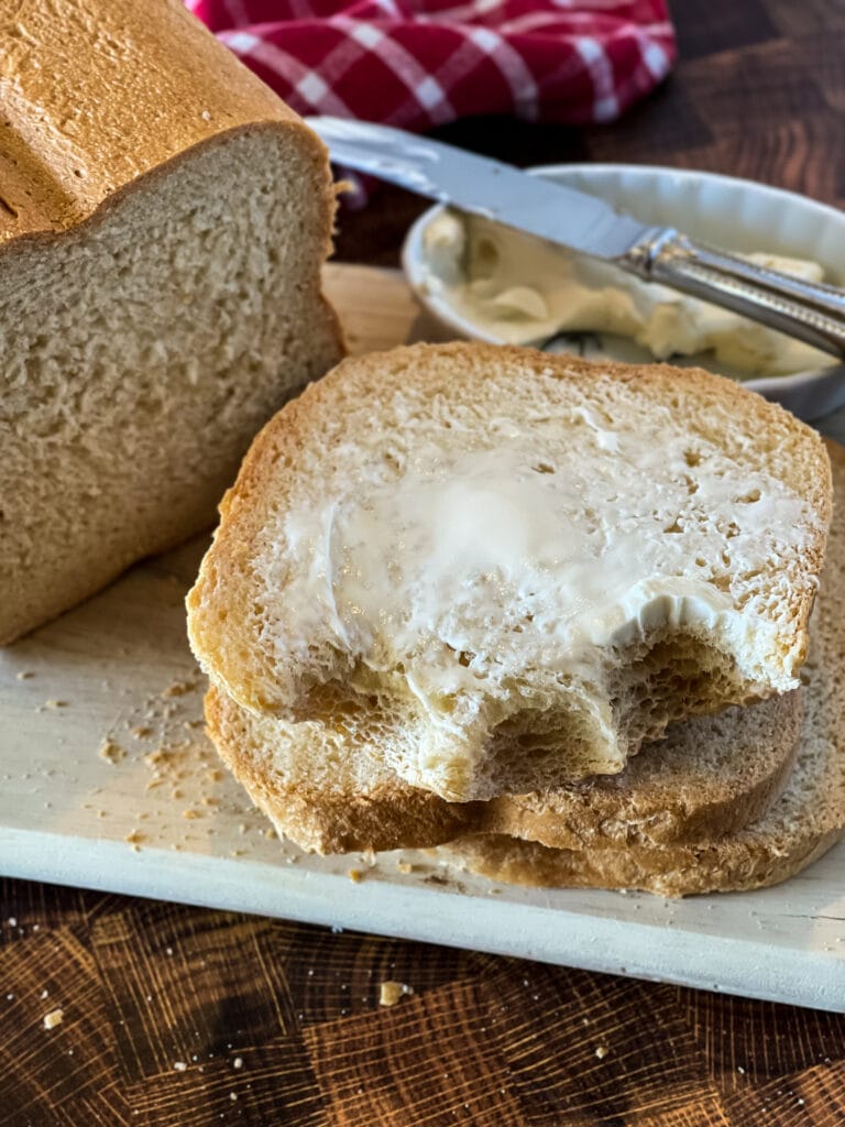 butter slice of white bread with bites out of it with butter dish and knive in background with partial loaf and red checked napkin in background