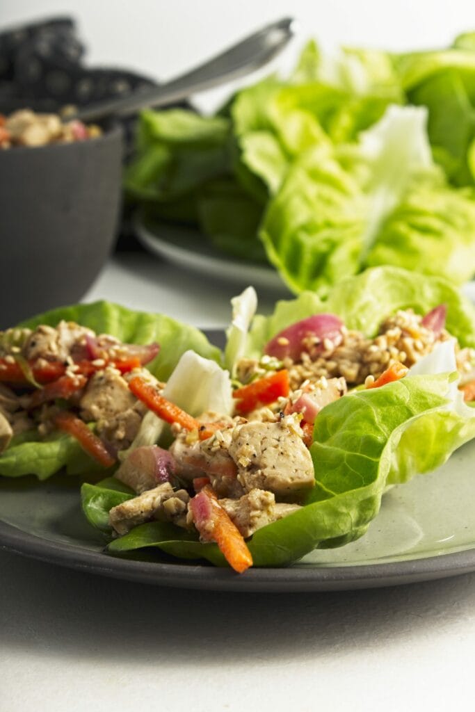 A small plate with three prepared tofu lettuce wraps with a bowl of the tofu mixture and a plate of butter lettuce in the background