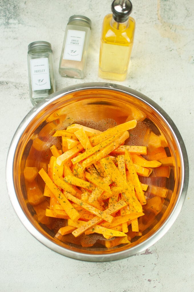 Seasoned and uncooked butternut squash fries in a metal bowl with the olive oil and seasonings behind it.
