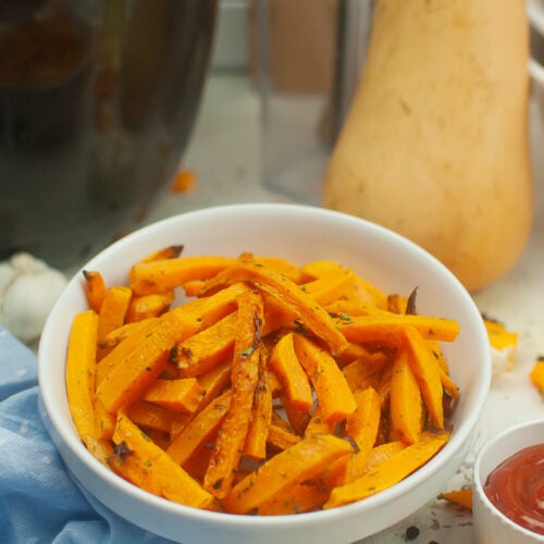 A white bowl of butternut squash fries with a butternut squash in the background with a bowl of ketchup on the side.