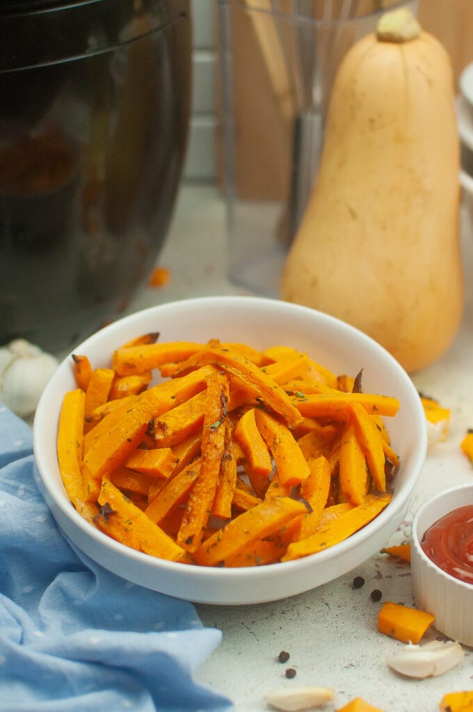 A white bowl of butternut squash fries with a butternut squash in the background with a bowl of ketchup on the side.