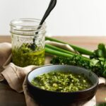 Chimichurri sauce in black bowl in foreground and in a jar in the background. Cilantro and parsley are on the side.