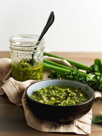 Chimichurri sauce in black bowl in foreground and in a jar in the background. Cilantro and parsley are on the side.