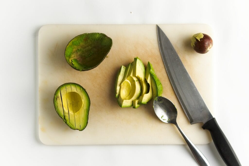 On the left, a avocado cut in half. The half on the top having been already sliced. The half on the bottom with slices but not yet scooped out. To the immediate right is the scooped out sliced avocado. Next to it is the spoon that scooped it and the knife. Finally, an avocado pit on the far right.