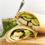 Two tofu wraps in lavish bread and wrapped in parchment paper on a wooden cutting board with parsley and chimichurri sauce in the background.