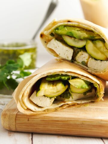 Two tofu wraps in lavish bread and wrapped in parchment paper on a wooden cutting board with parsley and chimichurri sauce in the background.