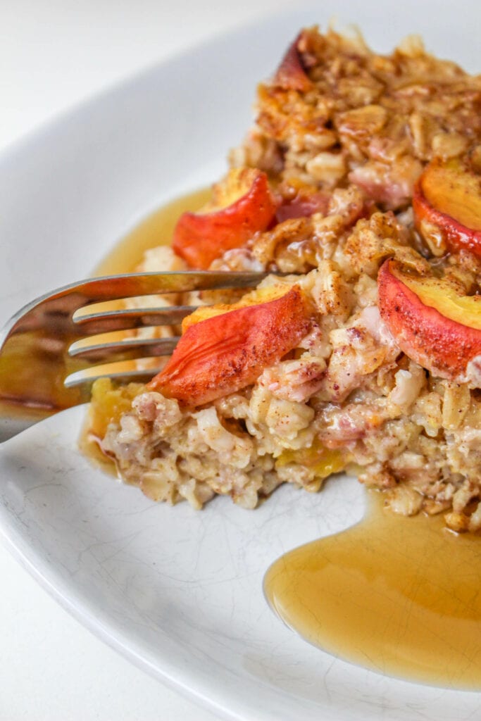 Taking a forkful of peach baked oatmeal from the slice on a white dish with maple syrup.