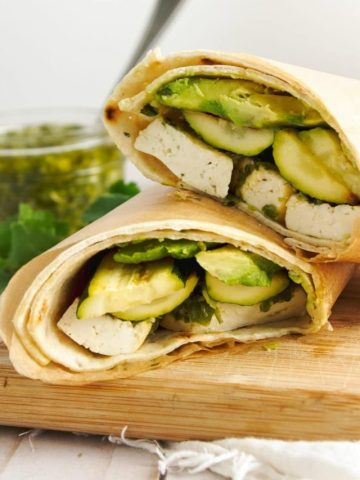Two tofu wraps in lavish bread and wrapped in parchment paper on a wooden cutting board with parsley and chimichurri sauce in the background.