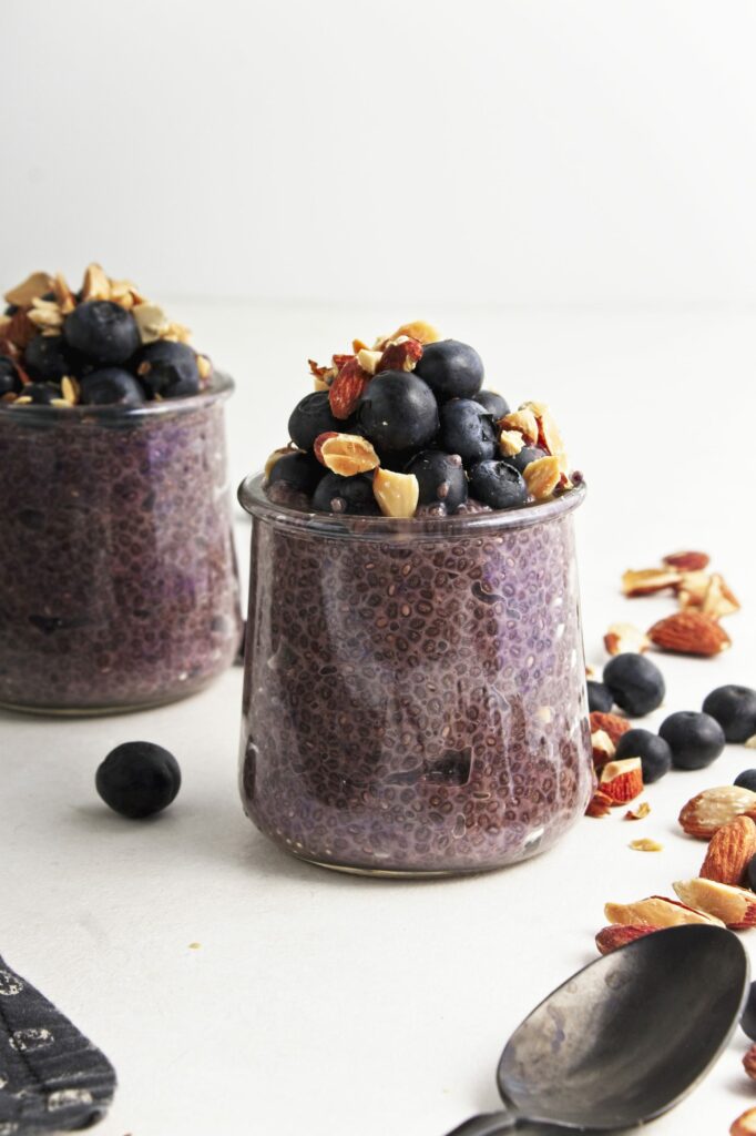 Two small glass jars with blueberry chia pudding and a mix of blueberries and chopped almonds on top with a scattering of chopped almonds and blueberries surrounding. With a dark spoon in the foreground.