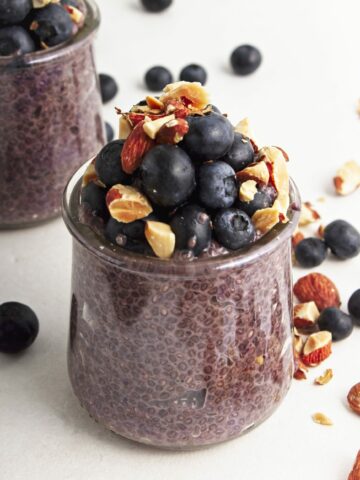 Two small glass jars with blueberry chia pudding and a mix of blueberries and chopped almonds on top with a scattering of chopped almonds and blueberries surrounding.