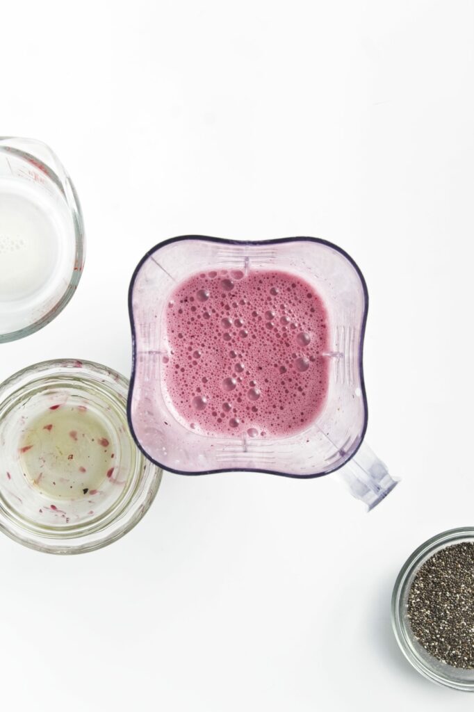 A blender with the blueberry mixture all blended in a blender surrounded a glass measuring cup, empty glass bowl with blueberry bits on the bottom, and a glass bowl with chia seeds.