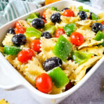 White rectangular serving dish containing pasta salad with a green striped napkin and jar of Italian dressing above and dried bow tie pasta at the bottom.
