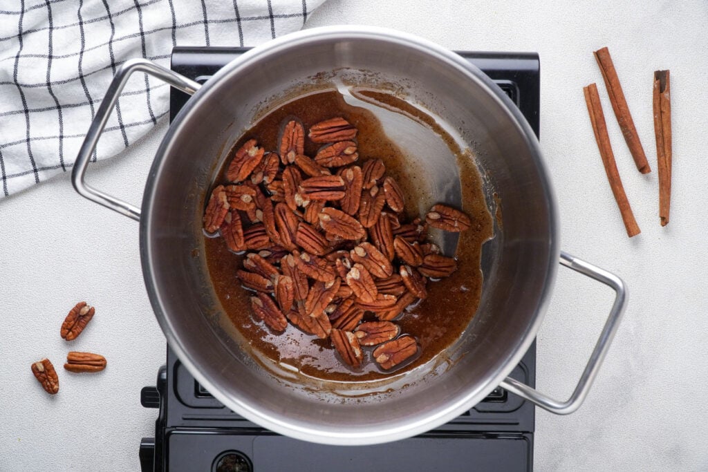 Adding the pecans to the melted ingredients in the pan with pecans, cloth, and cinnamon sticks on the sides and back.