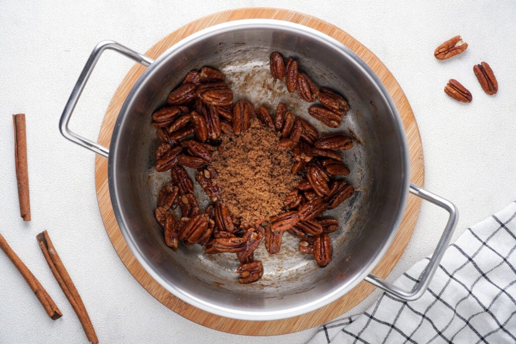 Adding in the extra brown sugar to the cooked pecans with cinnamon sticks, pecans, and cloth in fore and backgrounds.