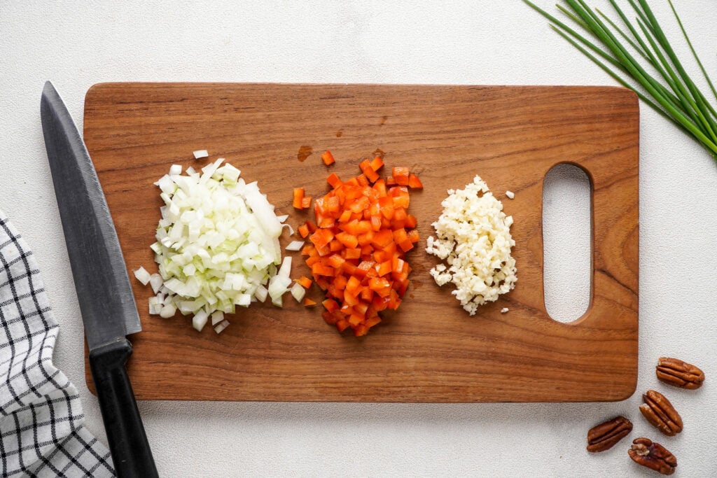 Chopped onion, red bell pepper, and garlic on a wooden cutting board. A cloth, knife, pecans, and chives surrounding.