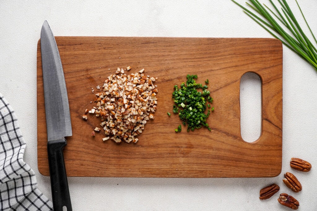 Chopped pecans and chives on a wooden cutting board. A cloth, knife, pecans, and chives surrounding.