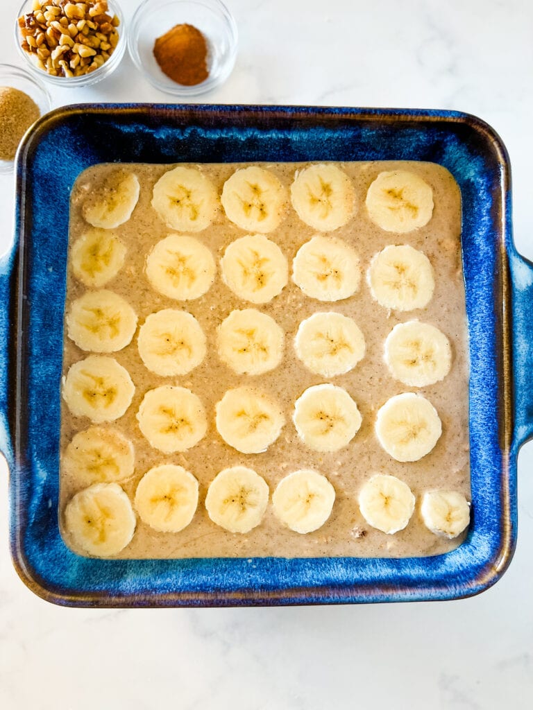 Adding the banana slices to the uncooked oats in the blue baking dish. Small bowls of brown sugar, chopped walnuts, and cinnamon are in the background.