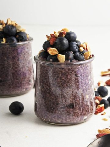 Two small glass jars with blueberry chia pudding and a mix of blueberries and chopped almonds on top with a scattering of chopped almonds and blueberries surrounding. With a dark spoon in the foreground.
