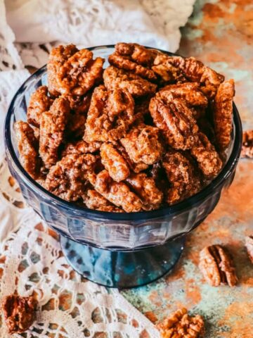 A blue glass candy dish with candied pecans on a lace napkin and a few pecans surrounding the dish.