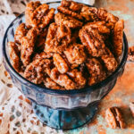 A blue glass candy dish with candied pecans on a lace napkin and a few pecans surrounding the dish.