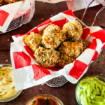 Pieces of popcorn tofu chicken in a wire basket with a red checked paper insert with two more in the background and three bowls of various dips in the foreground.