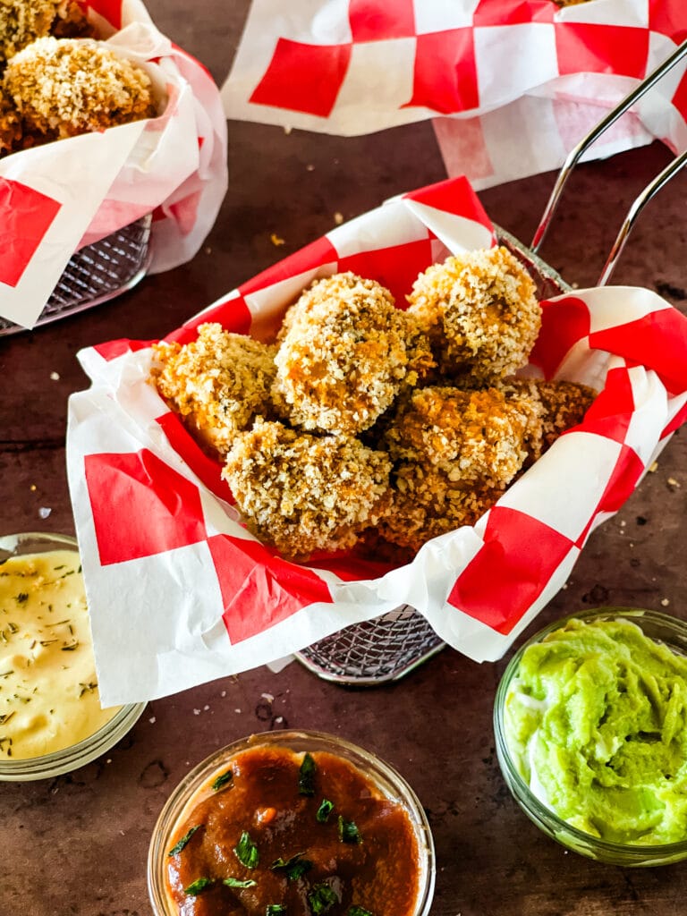 Pieces of popcorn tofu chicken in a wire basket with a red checked paper insert with two more in the background and three bowls of various dips in the foreground.
