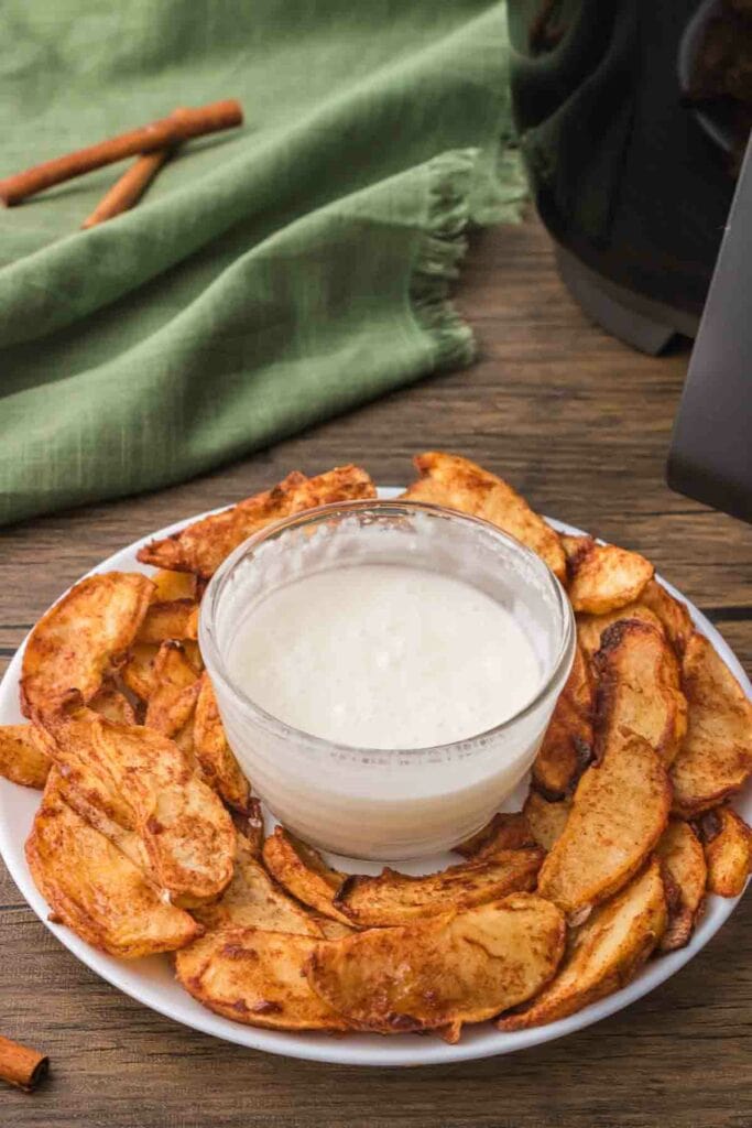 Air fried apple slices on a white plate with sweet coconut yogurt in a glass bowl in the center. A green napkin with two cinnamon sticks in the background.