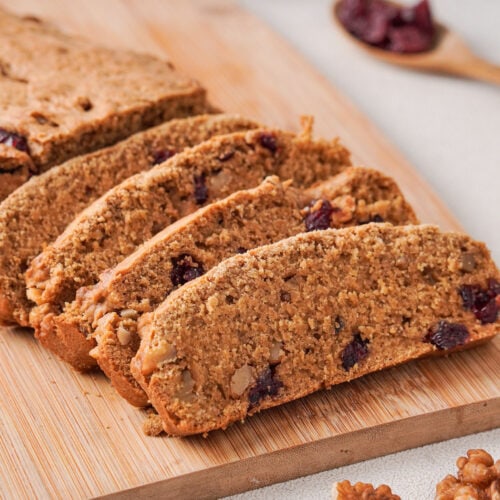 Slices of cranberry walnut loaf on a wooden cutting board. A wooden spoon with dried cranberries to the right. A blue striped cloth near the top. Three walnut halves near the bottom.