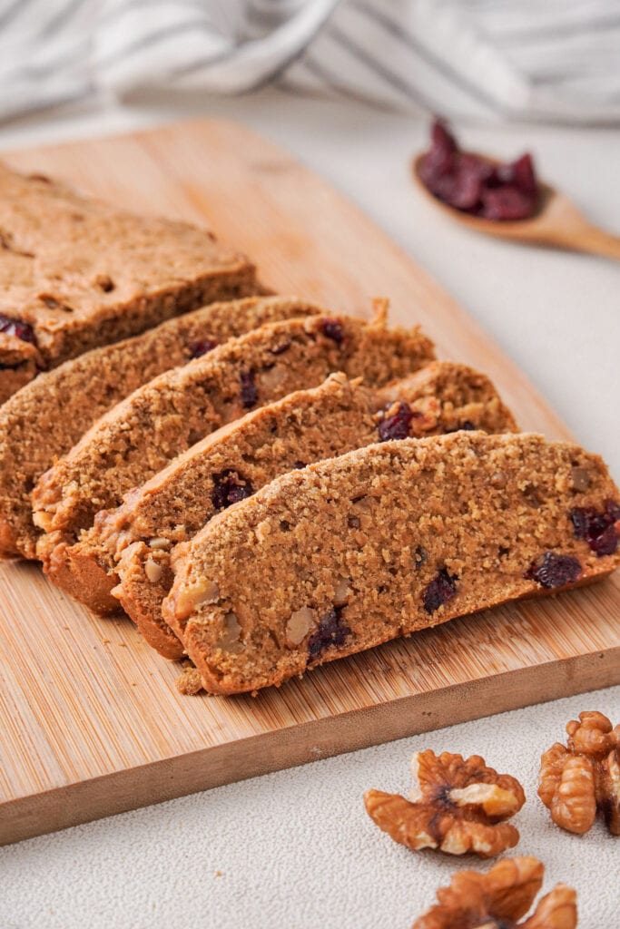 Slices of cranberry walnut loaf on a wooden cutting board. A wooden spoon with dried cranberries to the right. A blue striped cloth near the top. Three walnut halves near the bottom.