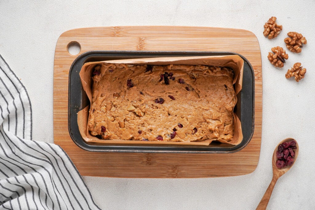 A loaf pan on a bread board with the cranberry walnut bread dough ready to bake with a blue striped cloth to the left, a wooden spoon on dried cranberries at the bottom right corner and four walnut halves on the top right corner.