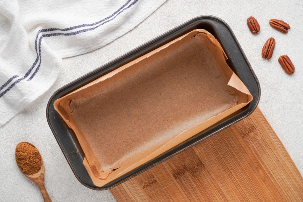 A loaf pan with parchment paper on a wooden cutting board with a wooden spoon with pumpkin pie spice and four pecan halves to the sides and a blue stripe cloth at the top.