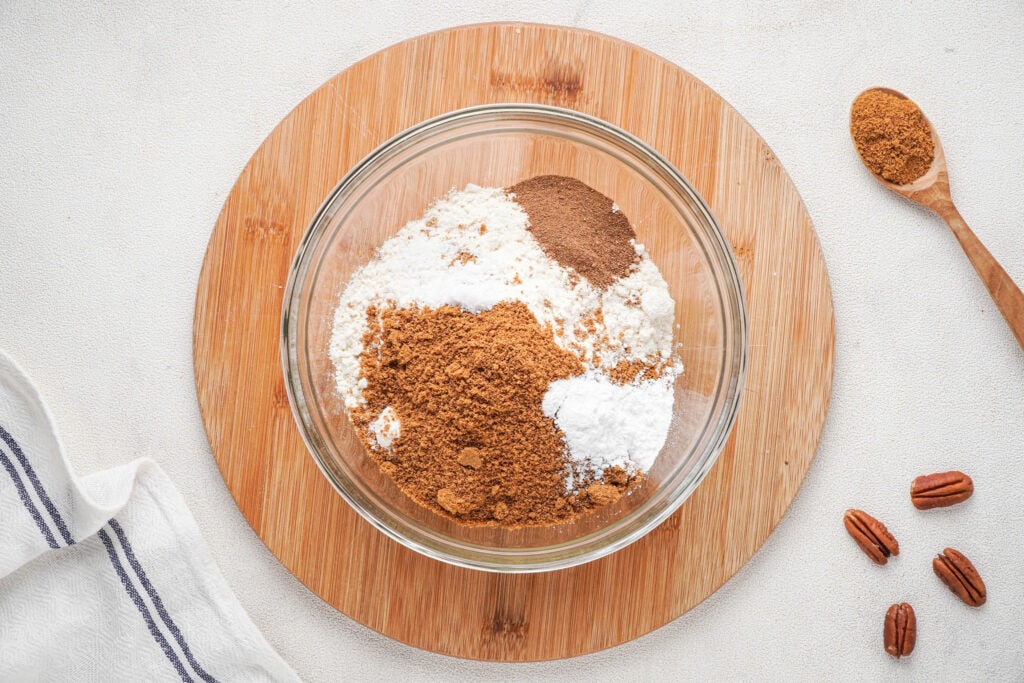 Dry ingredients in a glass bowl on a wooden round board with four pecans and a wooden spoon with pumpkin pie spice to the right.